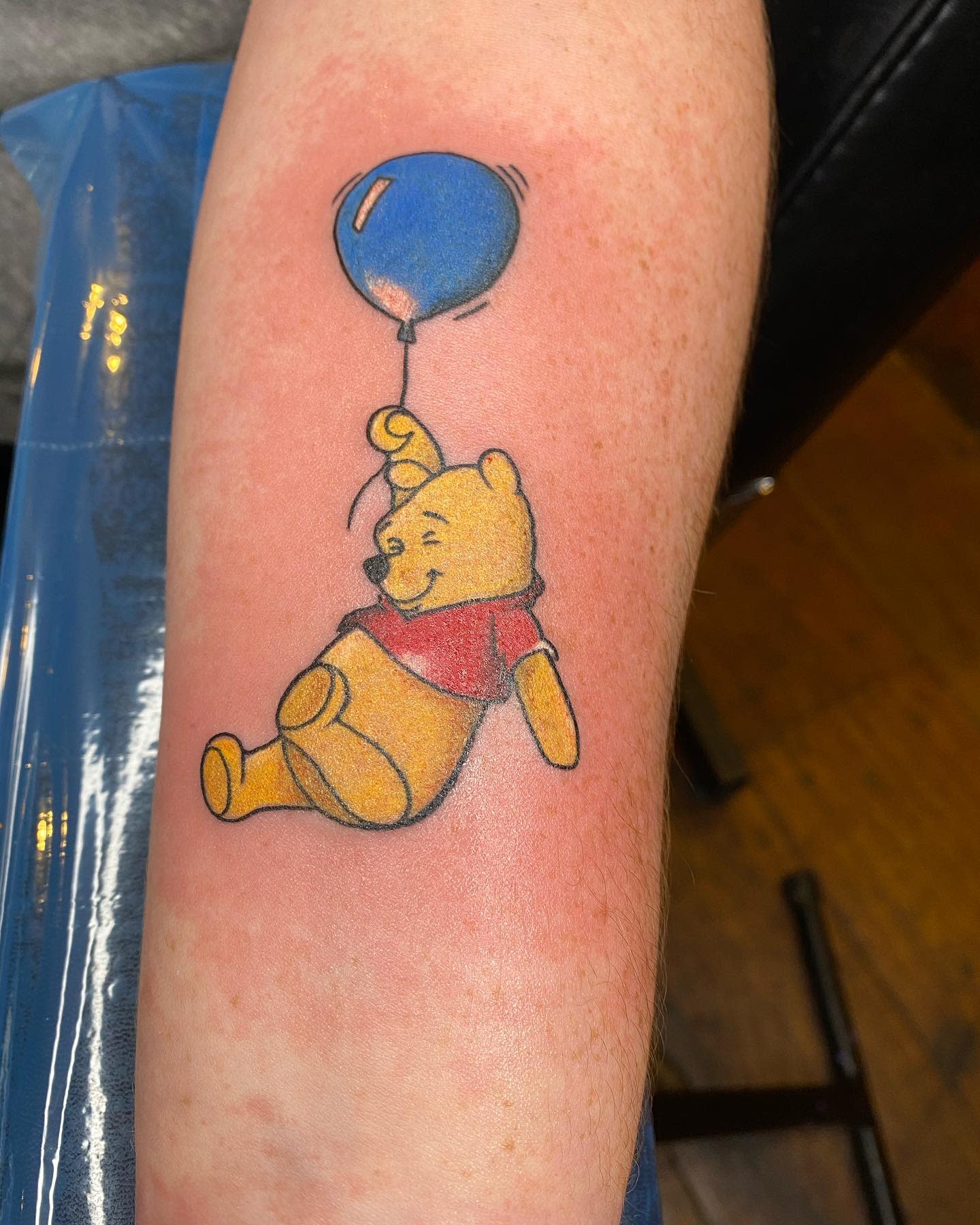 Color tattoo of Winnie the Pooh floating with a blue balloon on a person's forearm
